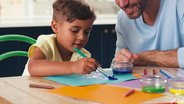 Mature father in kitchen at home with son sitting at table painting picture together - shot in slow motion