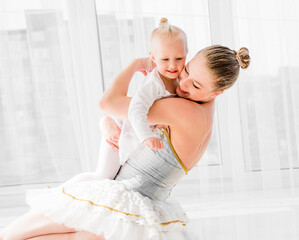 Young mother in swan ballet dress hugging little smiling daughter sitting on the floor in light...