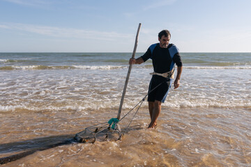 Mature fisherman dragging fishing rake while gathering clam shells in sea