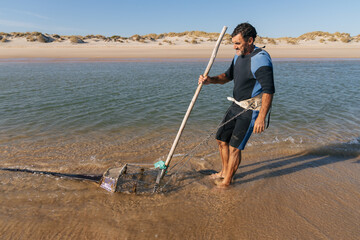Mature fisherman dragging fishing rake while gathering clam shells in sea