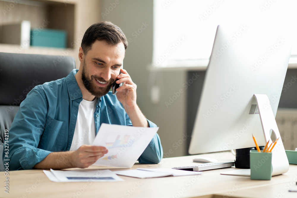 Poster smiling millennial businessman talking on cellphone and working with documents in office
