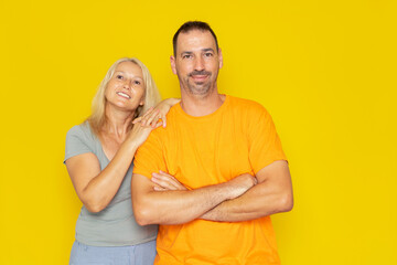 Caucasian real couple in their 40s posing happily, the man has his arms crossed and smiles as the woman leans on his shoulder. Isolated on yellow studio background.