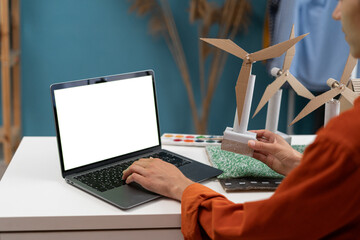 Young woman architect, working on an ecological construction project in her home office using laptop computer with white screen. Models of wind turbines at the foreground.