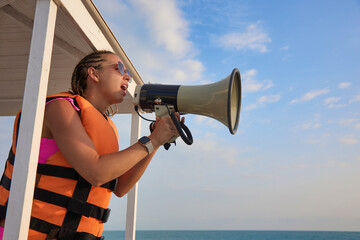 Young female lifeguard in an orange life jacket shouts an announcement into the loudspeaker for...