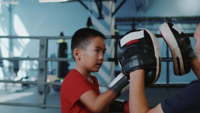 Young asian boy in boxing gloves intensively punching the boxing paws of his trainer and dodging his attacks on the ring close up. Child sports training in an indoor gym