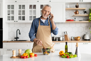 Smiling senior man talking on cellphone while cooking in kitchen interior