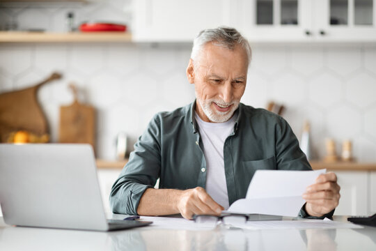 Happy Senior Man Reading Letter While Sitting At Desk In Kitchen