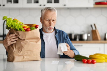 Prices For Food. Shocked Senior Man Checking Grocery Bills In Kitchen