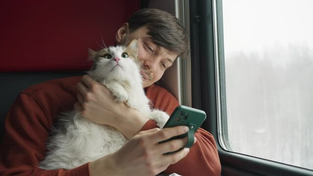 Joyful man looking at photos on his phone while hugging his cat on the train
