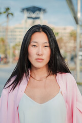 vertical individual portrait of serious asian young adult woman with toughtful expression. Teenage chinese girl standing outdoors with pensive attitude. One formal brunette lady looking at camera