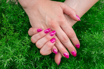 Close-up of well-groomed female nails with purple color gel. Beautiful clear fingers of a young girl with nice manicure and Glitter Nail Polish. Girl hands immersed in nature on a green background.