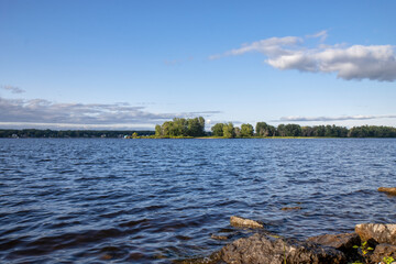 View of a forested island on the Ottawa River seen from the shoreline, rocks in foreground, daytime, blue sky, nobody