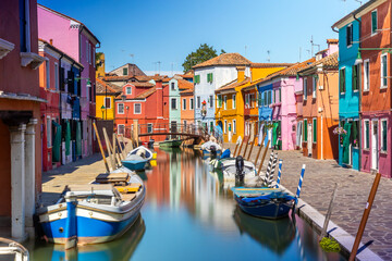 Colourful houses and buildings with reflection in blurred long exposure water of the river. Dock with moored boats on glassy water. Mirror of colors and bright vivid blue sky. Burano, Venice, Italy - obrazy, fototapety, plakaty