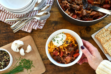 No bean keto beef and bacon chili stew being served with sour cream, cheese, chives by female in...