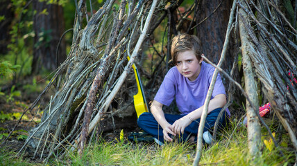 a happy 10 year old boy sits in a makeshift hut made of branches. joyful child spends summer holidays in a camp in nature © Ruslan Russland