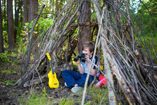 10 year old boy looks through binoculars, sits in a hut made of branches in a summer camp. a happy child spends his summer holidays hiking, outdoors, in the forest. - Powered by Adobe