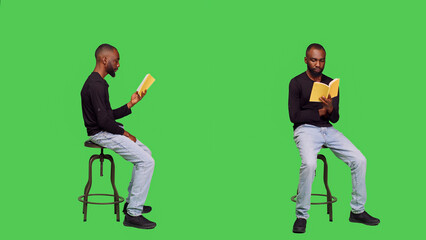 Fototapeta na wymiar Focused person reading story tale book in studio, sitting on chair over full body greenscreen. Young optimistic man enjoying novel lecture on camera, leisure activity with literature.