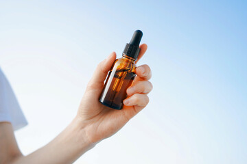 A young woman is holding a glass transparent jar of facial serum against a blue sky background....