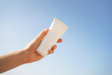 Female hand holding a white tube of sunscreen mockup on a blue sky background. Spa concept,...