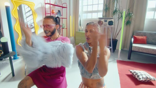 Artistic LGBTQ couple of cross-dressed guy and his gender fluid partner in y2k outfit performing energetic vogue dance, smiling and posing for camera in trendy designed studio. Zoom shot