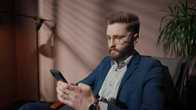Attractive Caucasian man with beard in stylish suit typing message on his smartphone. Comfortably sitting in armchair near lamp with bright light in shadow room. Using technology device at workplace.