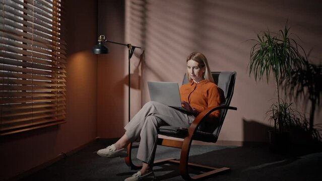 Zooming in on Caucasian female worker with blonde hair working in dark room alone. Actively typing text on her laptop. Concentrating on work task. Moderm office. Concept of technology.