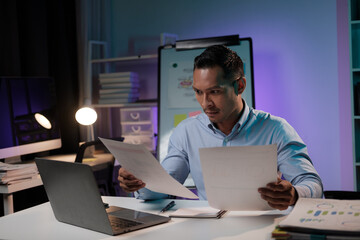 Hardworking Asian businessman focusing on his project, holding papers in his hands, working overtime at night alone in the office.