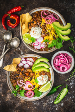Chili con carne in two bowls with garnishes, pickled red onions and spoons in blowls on dark background