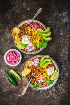 Chili con carne in two bowls with garnishes, pickled red onions and spoons in blowls on dark background