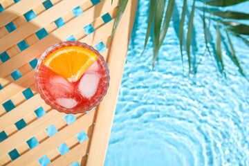 Glass of cold Negroni cocktail and palm leaf on wooden grid near swimming pool