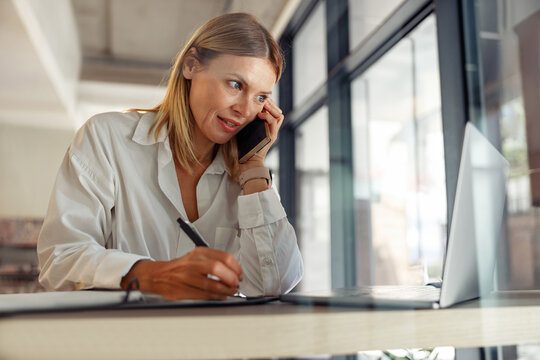 Woman entrepreneur making notes and talking by phone with client while sitting in coworking 