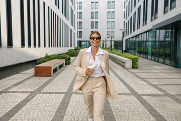 Smiling business woman with coffee walking on modern building background during break time