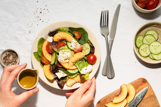 Hands drizzling jug of olive oil over mozzarella and nectarine salad in ceramic bowl, with ingredients surrounding.