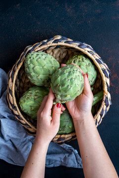 Woman Hands Taking A Fresh Artichoke From A Basket Filled With Artichokes Photographed From Top View.