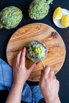 Woman hands halving an artichoke from the stem using a knife on a wooden cutting board.