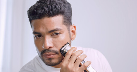 A man is using electric shaver to trim mustache and beard while standing in front of mirror in bathroom at home.