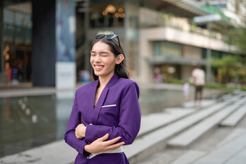 A lovely lady standing outside, in the city, in a purple suit and sunglasses holding her tummy with both hands from laughing and eyes are closed. Buildings and stairs are in the background.