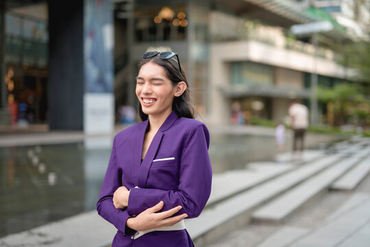 A lovely lady standing outside, in the city, in a purple suit and sunglasses holding her tummy with both hands from laughing and eyes are closed. Buildings and stairs are in the background.