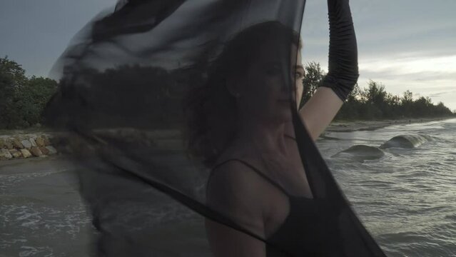 Young elegant woman in black dress dancing with hands up holding black scarf on the pier. Close up. Summertime, vacation, freedom concept