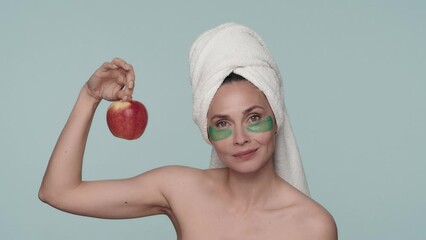 Seminude woman with a towel on her head and green hydrogel patches under her eyes on a blue background. A woman holds a ripe red apple in her hand and looks at it. Slow motion. - Powered by Adobe