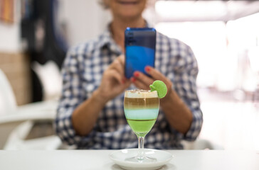 Senior woman using phone sitting at cafe table, a glass of local specialty of Canary island called barraquito in front to her, made with condensed milk, coffee and liqueur