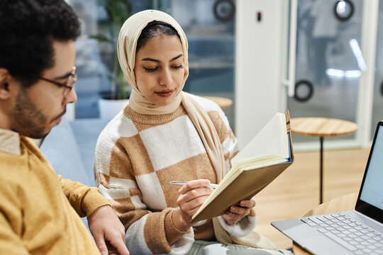 Portrait of young Muslim woman wearing head scarf in office and talking to male colleague discussing notes
