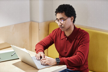 Portrait of Middle Eastern young man wearing glasses and using computer at desk in booth with electric lighting, copy space