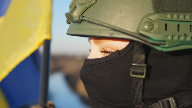 Gaze of ukrainian army woman in helmet and balaclava outdoor. Female soldier looks at sunset against a waving blue-yellow flag at countryside. Concept of peace and support against russian aggression