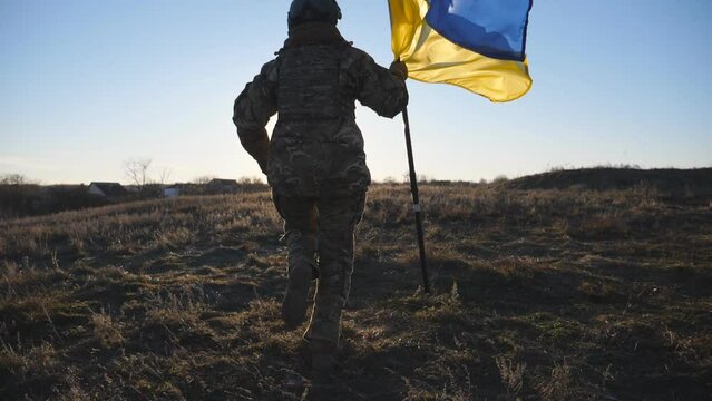 Young woman in camouflage uniform jogs with waving flag of Ukraine against sunset. Female soldier of ukrainian army runs with lifted national banner on the field. Invasion resistance. Rear view