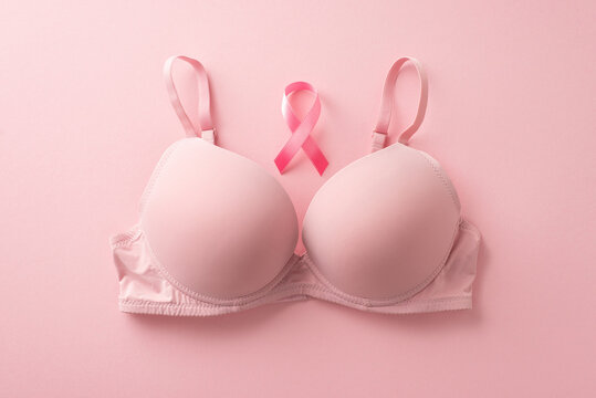 Symbolic breast cancer awareness. Pink-themed top view photo featuring a bra and pink ribbon on a pastel pink surface. Space available for text or advert placement