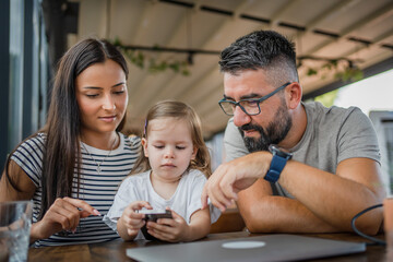One toddler girl sitting in cafe with mother and father and looking at mobile phone, modern parenting concept