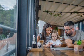 One toddler girl sitting in cafe with mother and father and looking at mobile phone, modern parenting concept