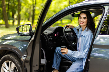 Beautiful businesswoman is sitting at steering wheel of her car. She is drinking coffee and smiling.