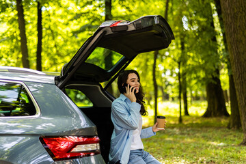 Young happy female sitting in car trunk and looking away while having phone conversation